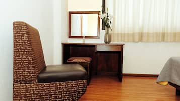 Una habitación con un sillón estampado, un escritorio pequeño con un espejo, un jarrón con flores y una cama con una manta.