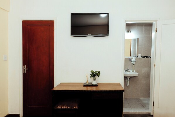 Una habitación con escritorio de madera, televisor de pantalla plana en la pared, una puerta y un baño con lavamanos y espejo.