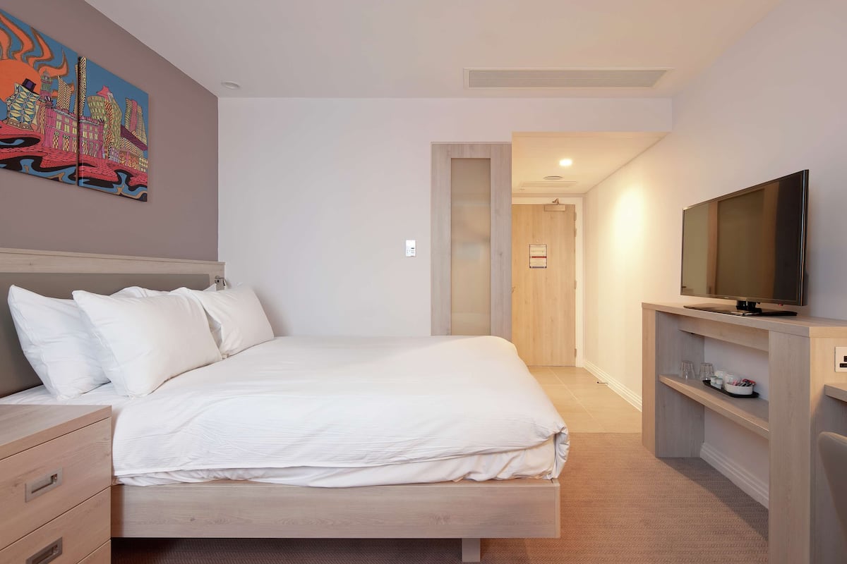 A modern hotel room with a bed, a TV, a wooden dresser, and a painting on the wall.