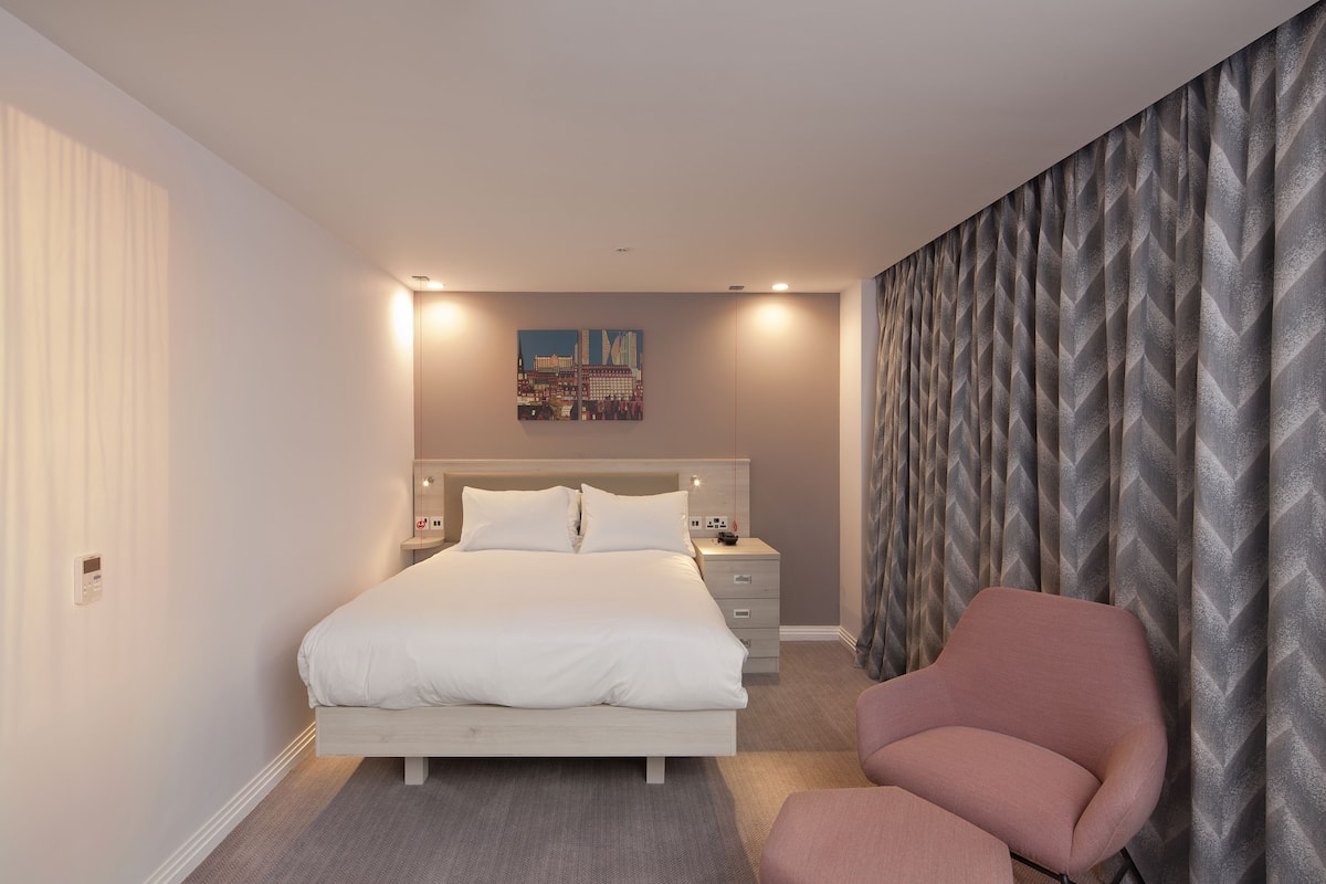 A bedroom with a bed, bedside tables, a chair, and a cityscape painting on the wall.