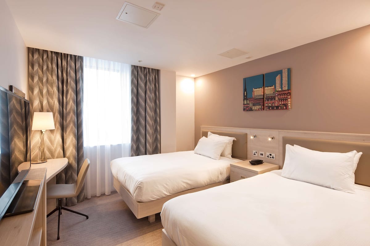 A hotel room with two beds, a desk, a lamp, and a painting on the wall.