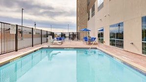 A swimming pool with lounge chairs and umbrellas, surrounded by a fence and a building with large windows. - Holiday Inn Express Kermit by IHG (Kermit)