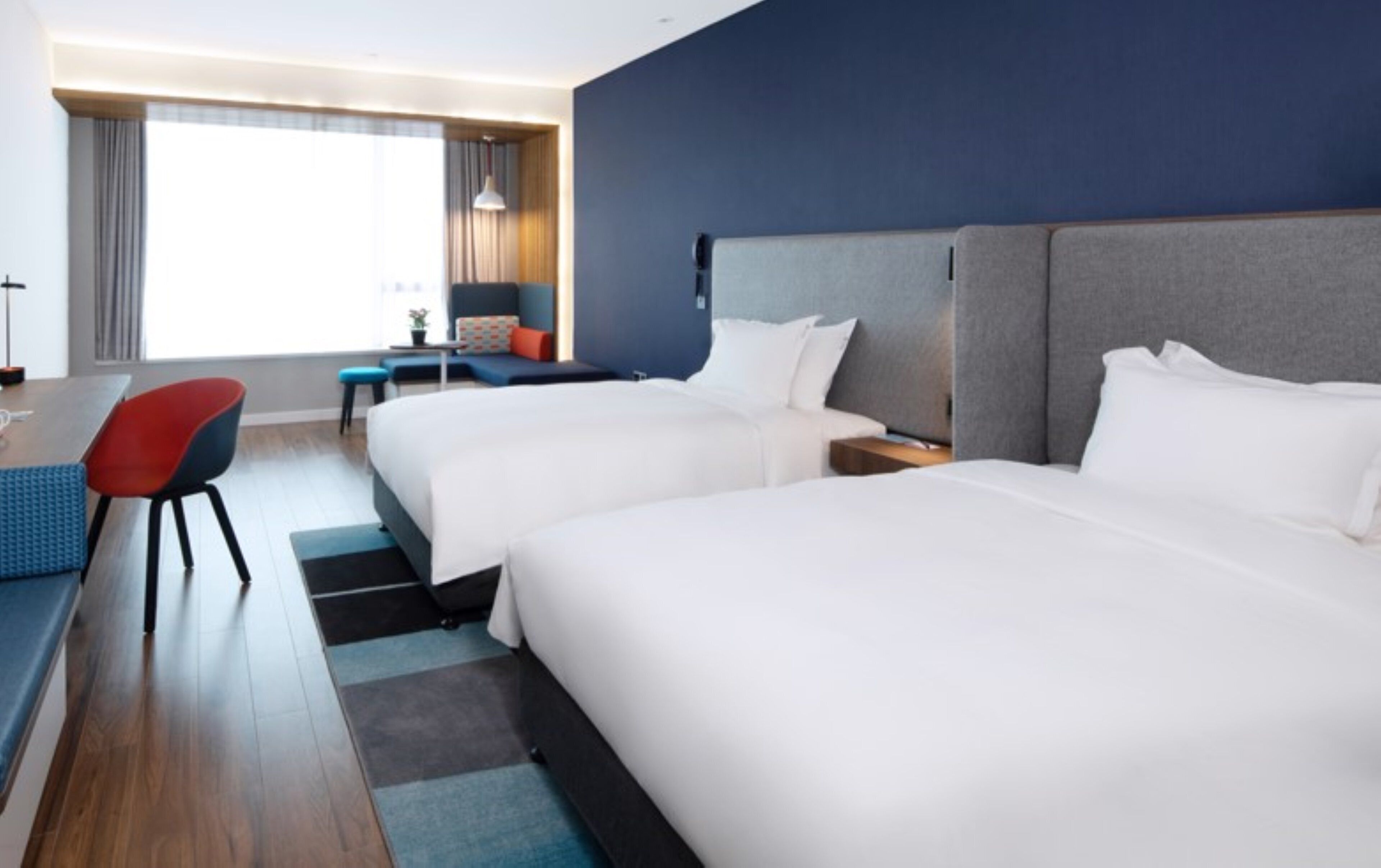 A hotel room with two beds, a desk, a chair, and a view of the city.