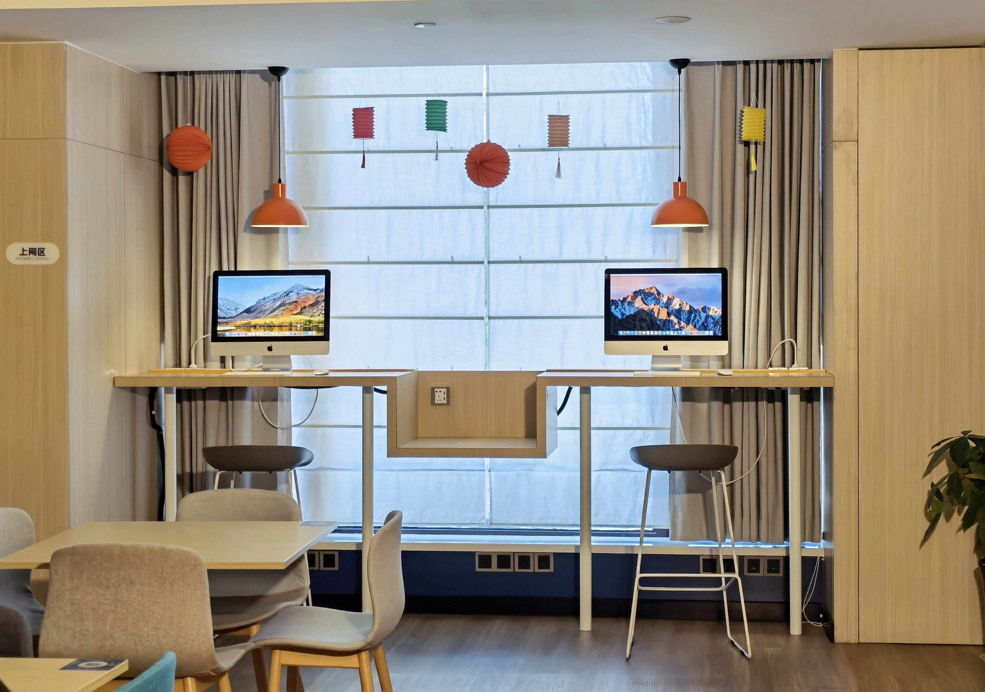 Una habitación de hotel moderna con dos escritorios, cada uno con un monitor de computadora, una silla y un taburete pequeño. La habitación tiene un ventanal con cortinas, una planta y un estante montado en la pared con objetos decorativos.