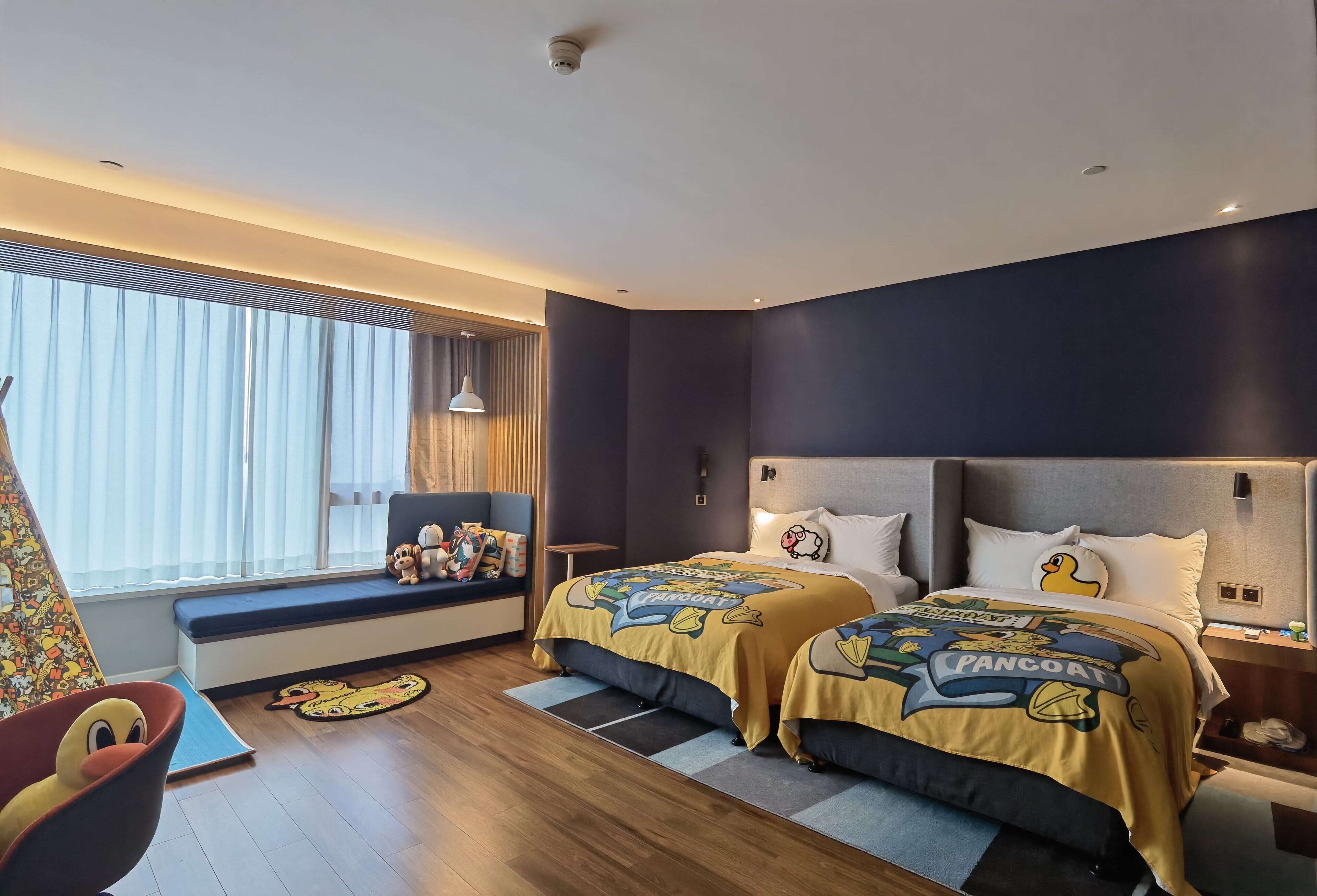 A hotel room with two beds, a large window with blinds, a sofa with a colorful cushion, and a small table with a lamp.
