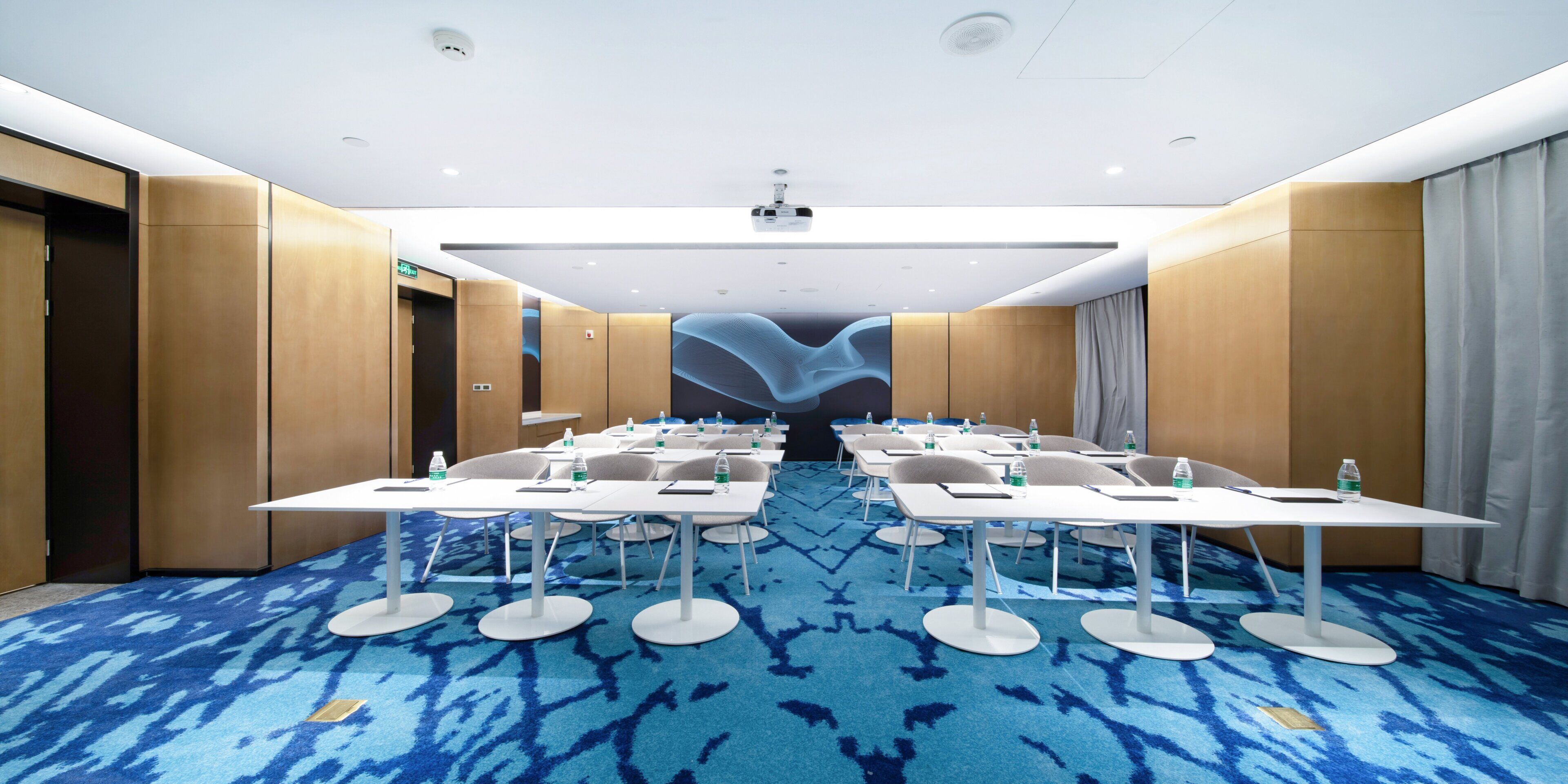 Un salón de conferencias con alfombra azul, mesas y sillas blancas, y botellas de agua.