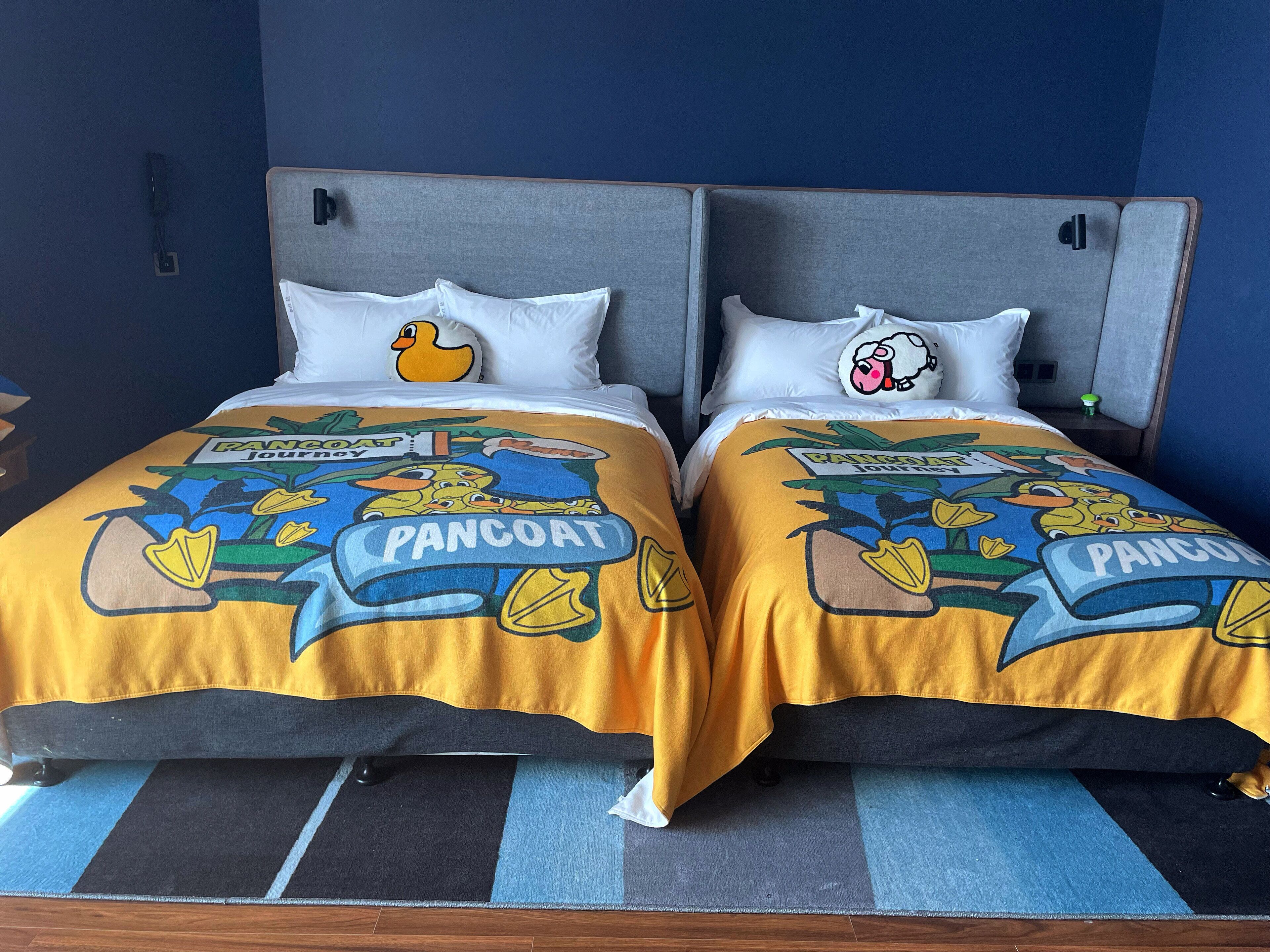 A double bed with a headboard, two pillows, and a bedspread featuring cartoon ducks and the word "PANCOAT."