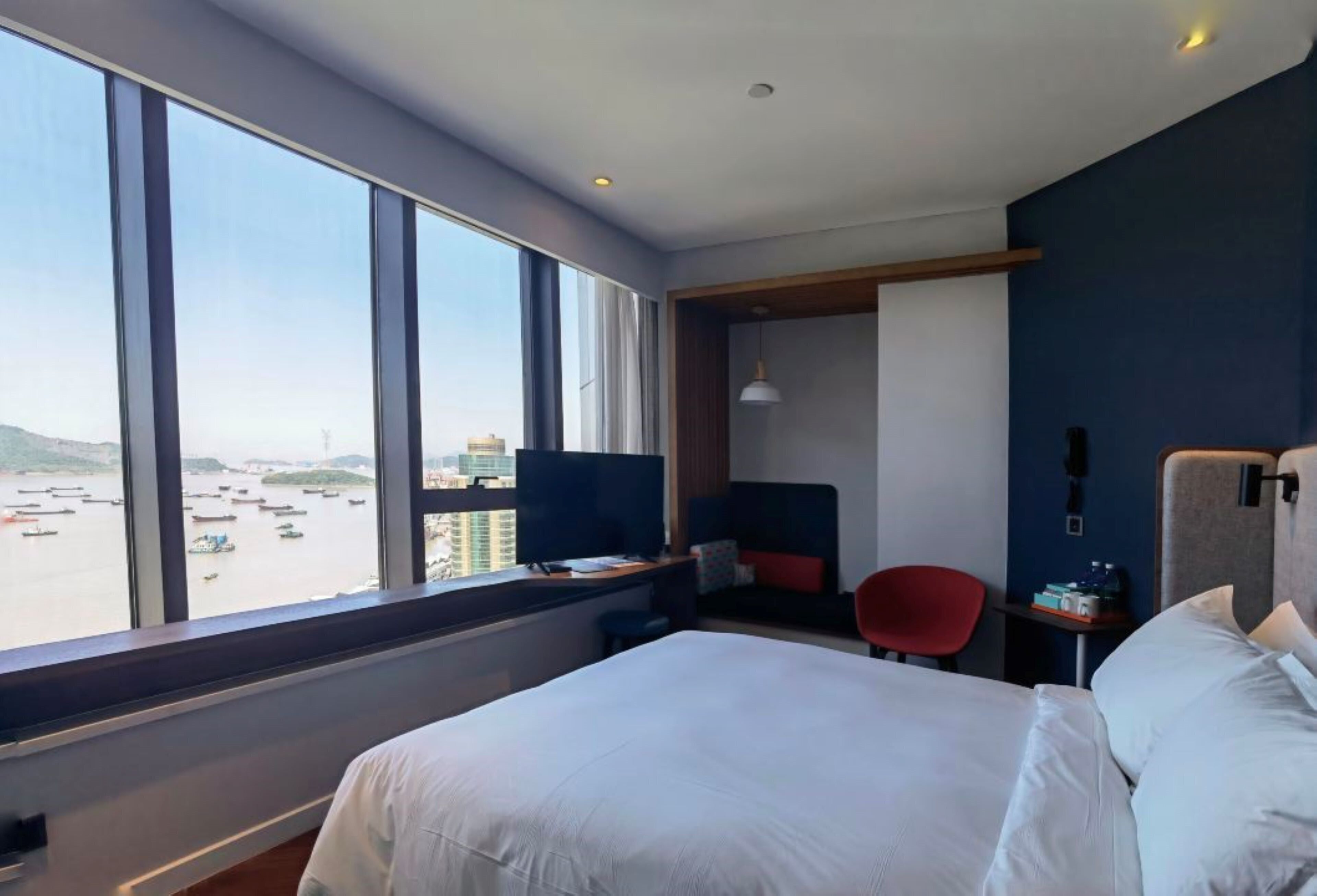 Hotelkamer met een groot bed, een bureau met een computer, een stoel en uitzicht op het water door grote ramen.