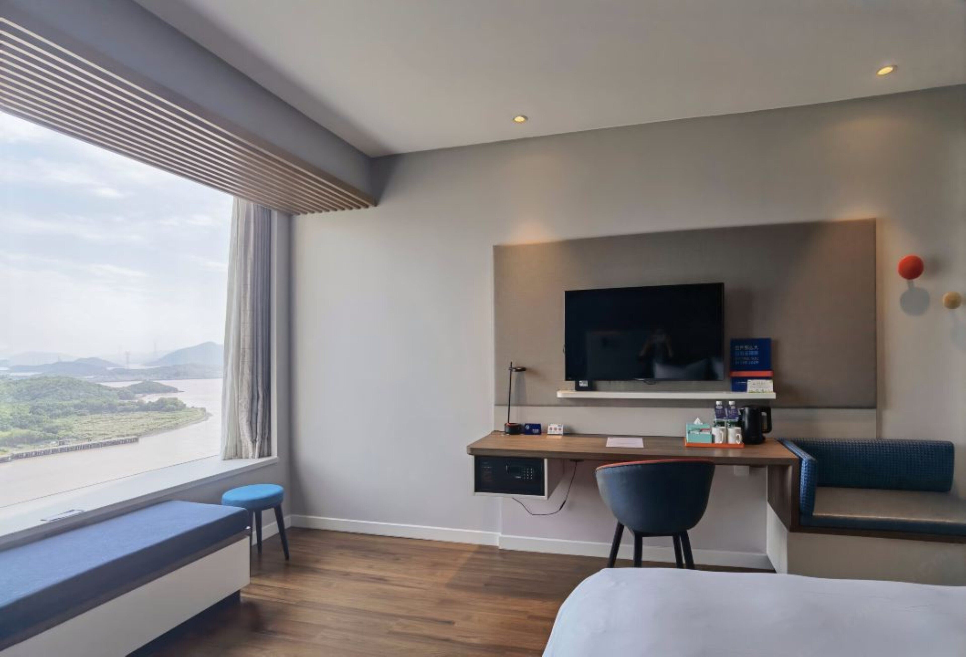 A hotel room with a large window offering a view of a river and mountains, a desk with a chair, a bed, and a TV mounted on the wall.