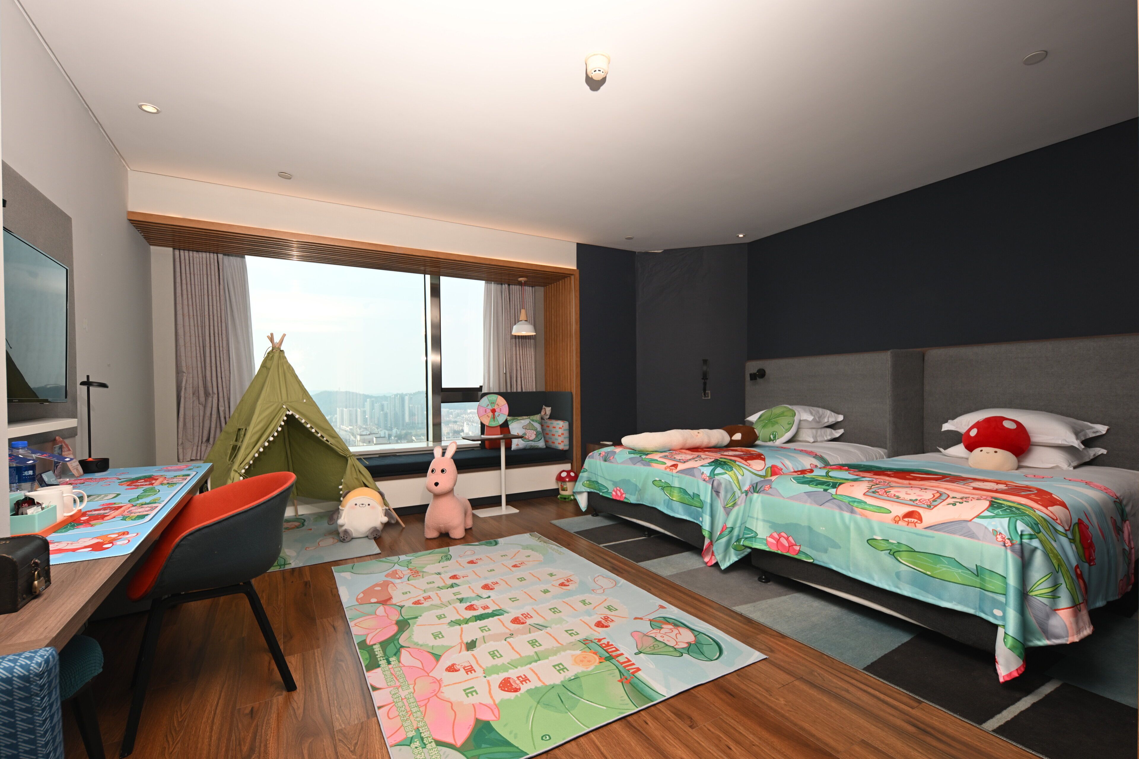 A modern hotel room with a large window, a bed with a colorful bedspread, a desk with a chair, and a small play area with a tent and toys.
