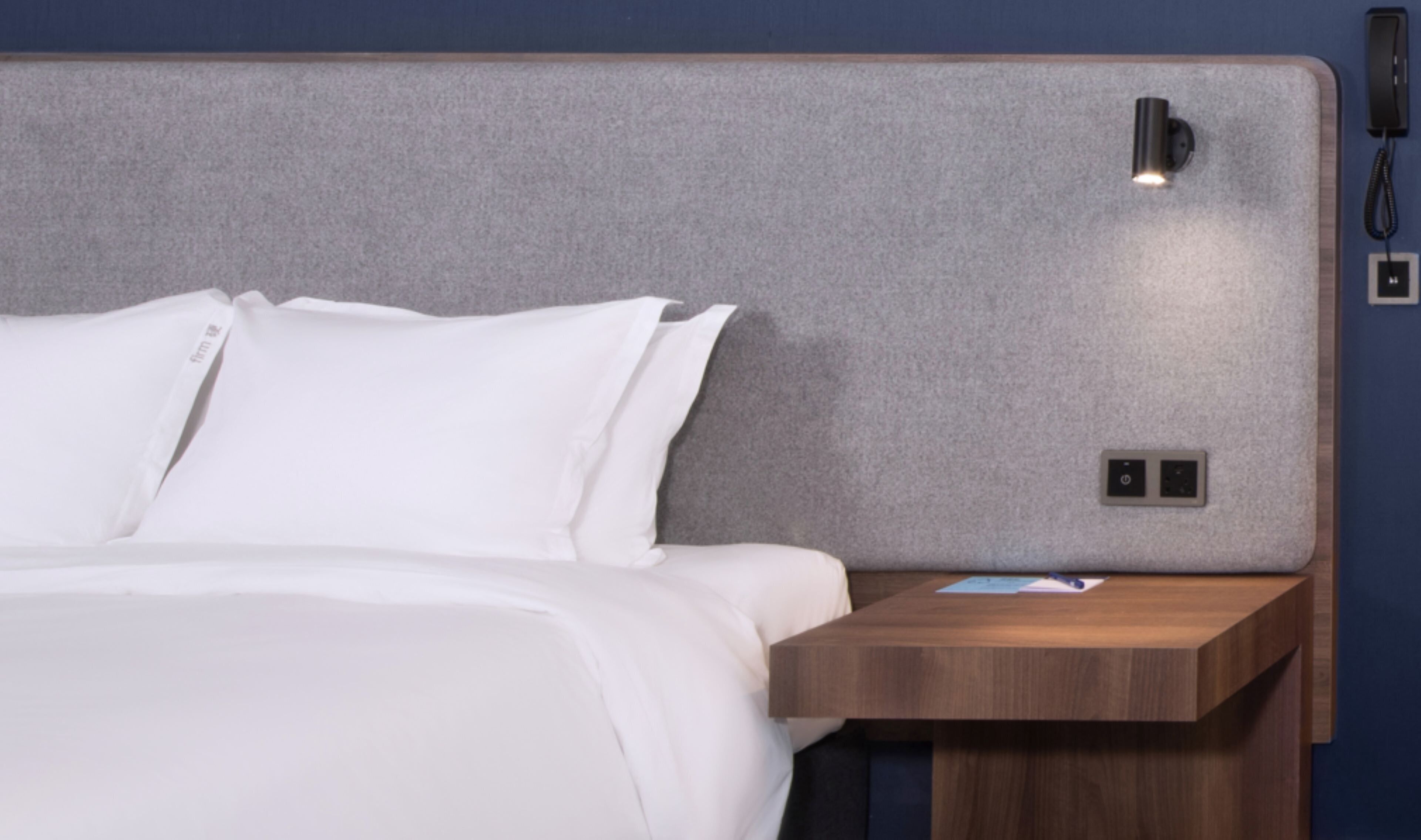 Un letto ben rifatto con lenzuola bianche, un comodino in legno con una lampada e una presa di corrente.