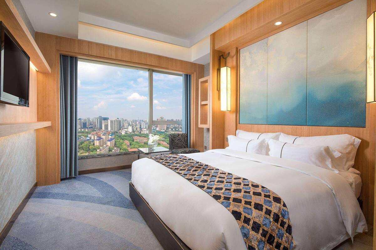 Habitación de hotel con una cama grande, televisor, vista a la ciudad y un cuadro en la pared.