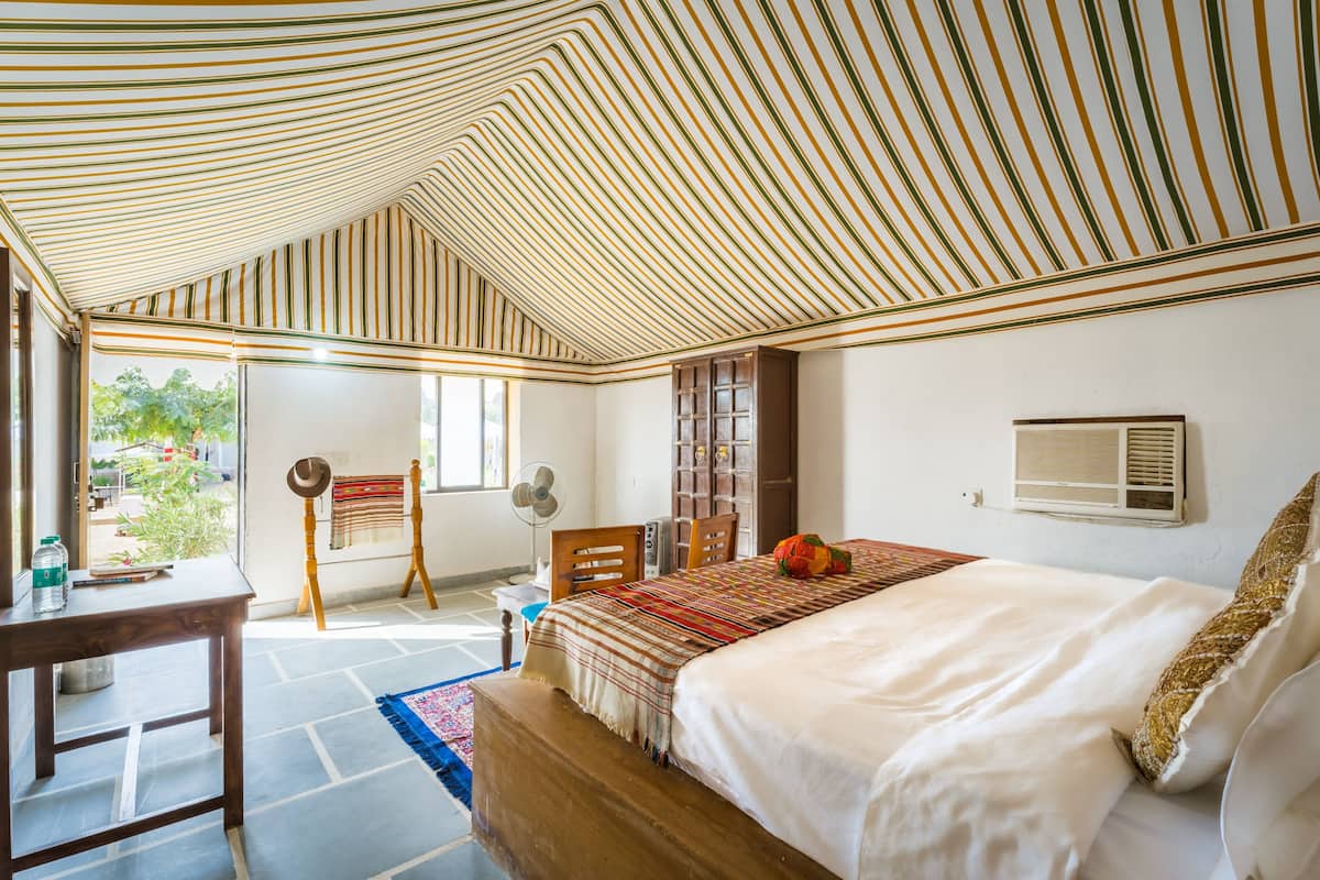 A bedroom with a striped ceiling, a bed with a white comforter, a wooden headboard, a small table with a water bottle, and a window with a view of greenery.