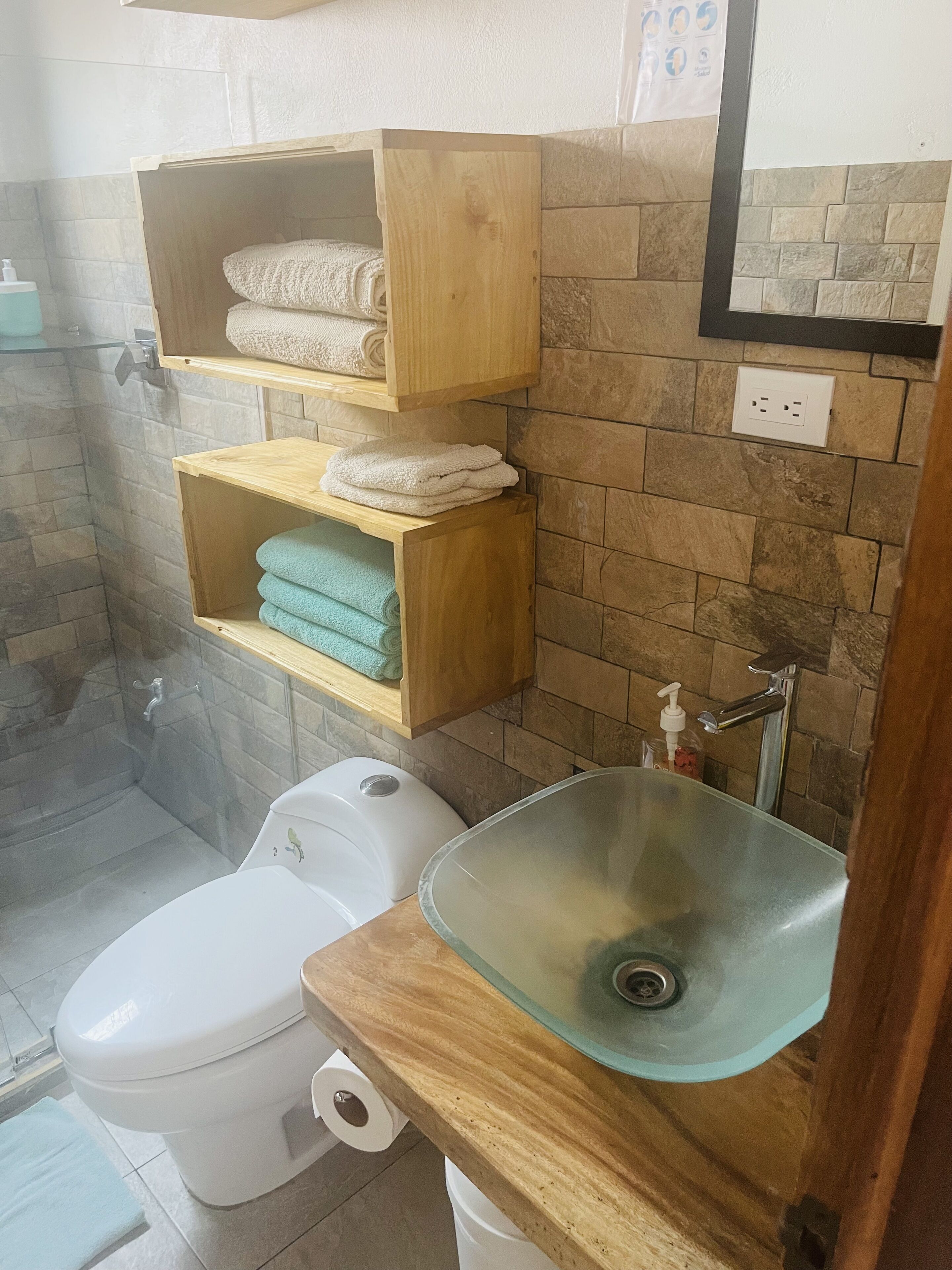 Un baño con inodoro, lavabo de cristal y repisas de madera con toallas.