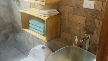 Un baño con inodoro, lavabo de cristal y repisas de madera con toallas.