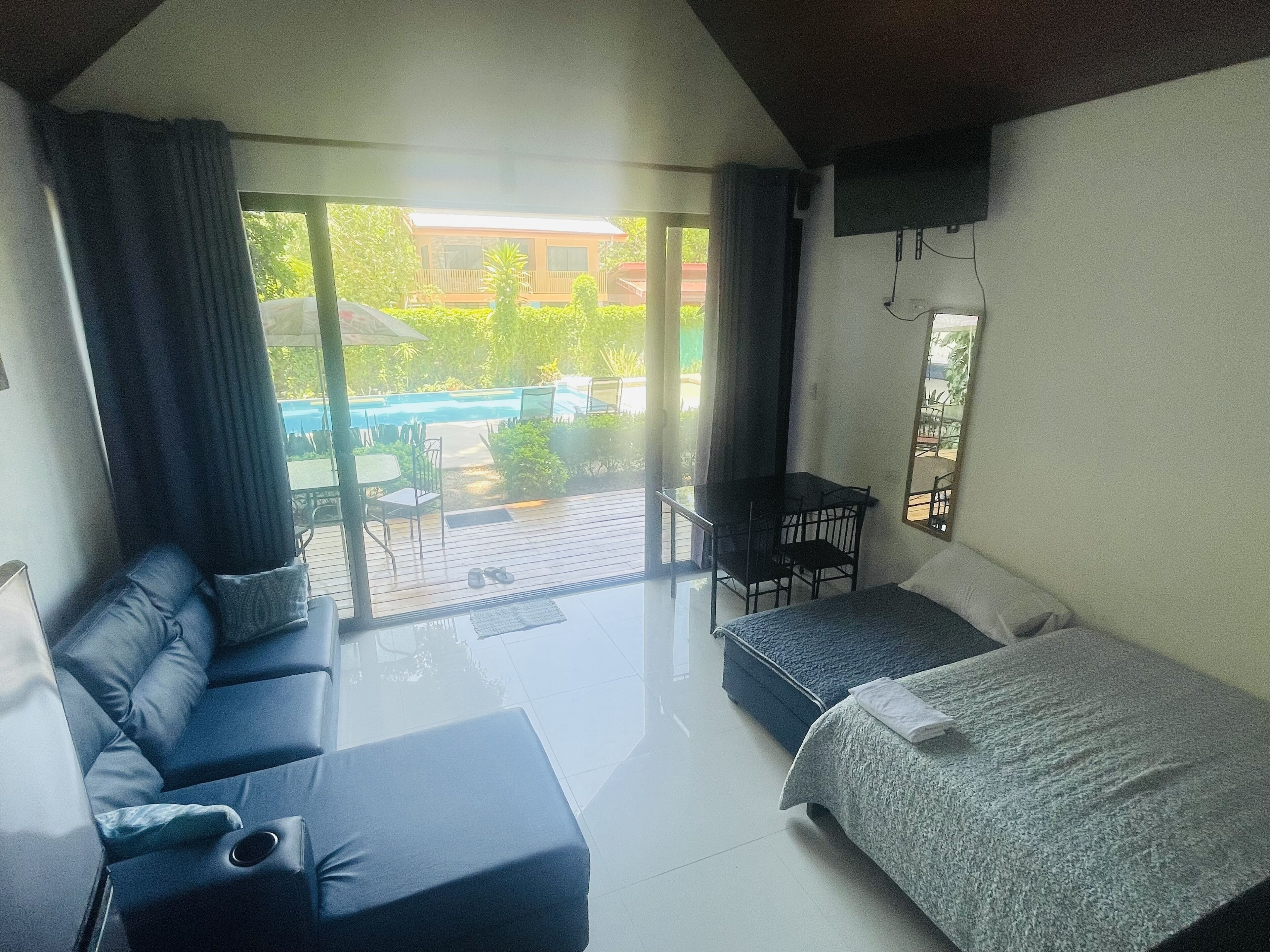 A modern room with a large bed, a sofa, and a small table. There is a sliding glass door leading to an outdoor area with a pool and patio furniture.