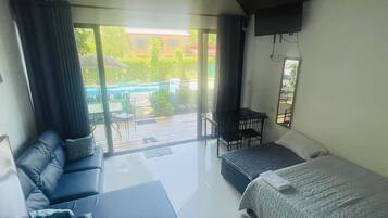A modern room with a large bed, a sofa, and a small table. There is a sliding glass door leading to an outdoor area with a pool and patio furniture.