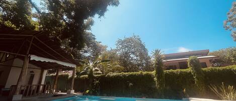A swimming pool with a tiled deck, surrounded by greenery and a building in the background.