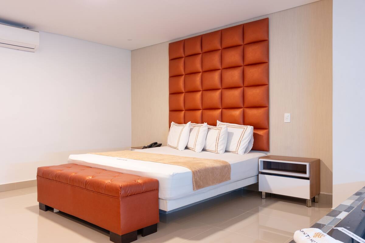 A modern bedroom with a large bed, an orange upholstered bench, a bedside table, and a wall-mounted air conditioner.