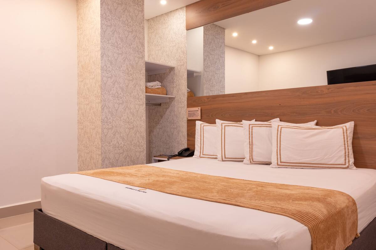 A modern hotel room with a wooden headboard, a large bed with white bedding and decorative pillows, a bedside table with a phone, and a built-in shelf with toiletries.