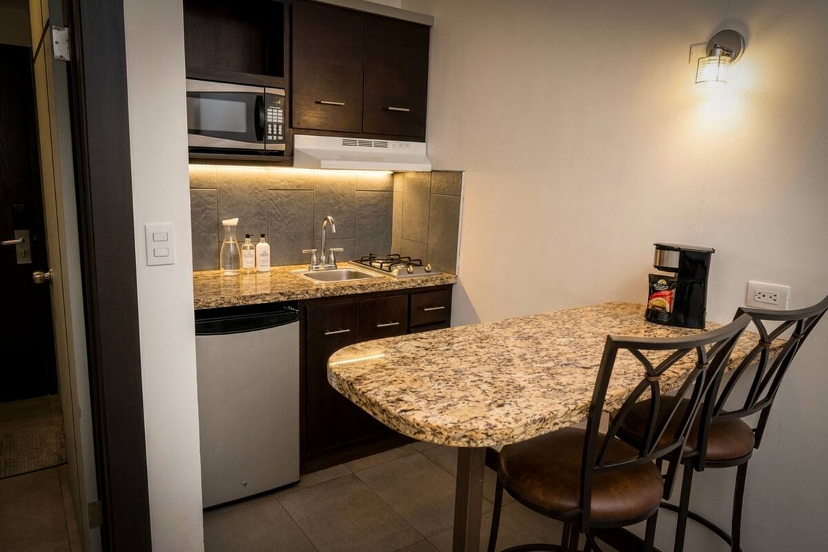 A compact kitchen with a built-in microwave, a gas stove, and a coffee maker. There is a granite countertop and two bar stools.