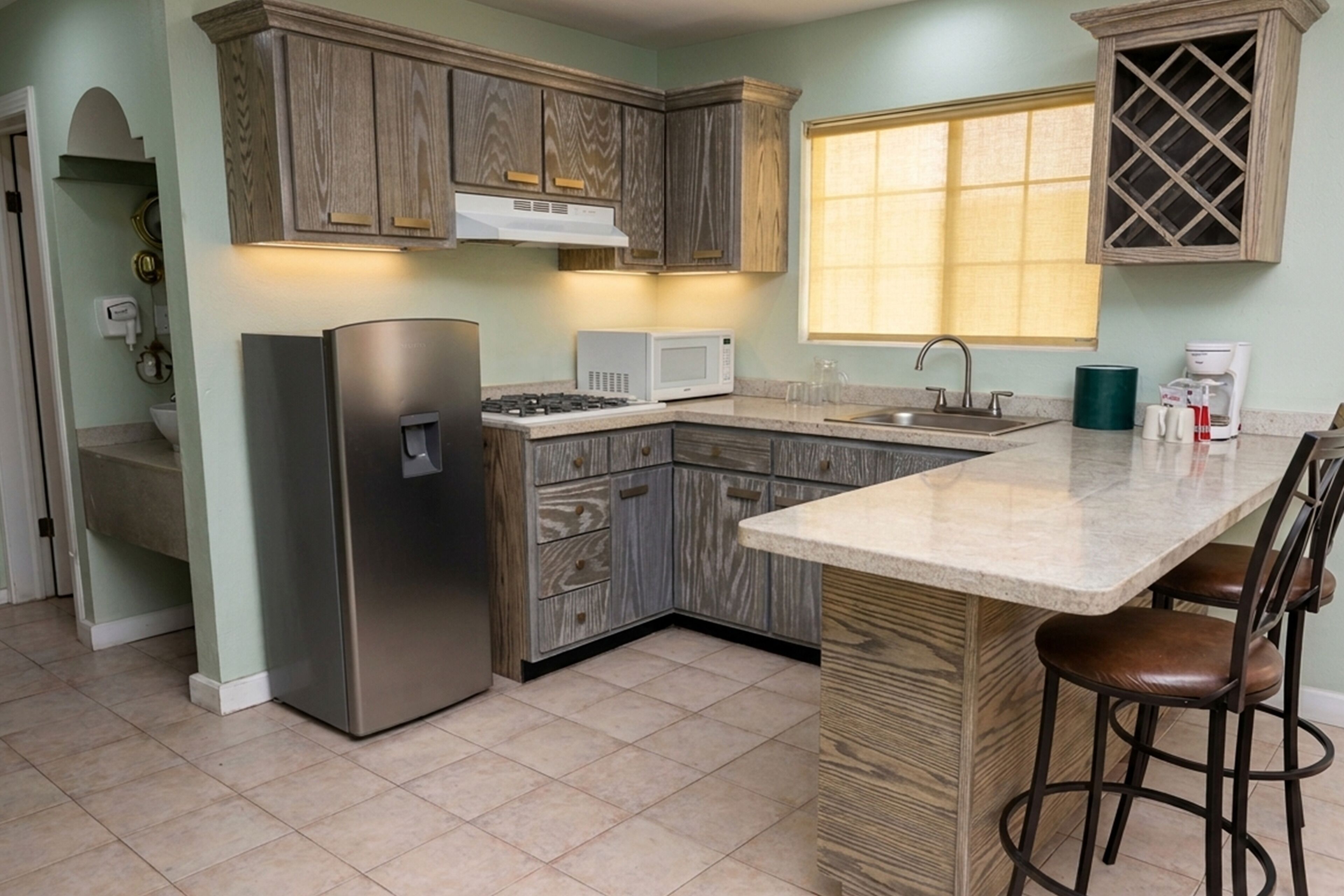 A kitchen with wooden cabinets, a stainless steel refrigerator, a microwave, a sink, and a bar area with stools.