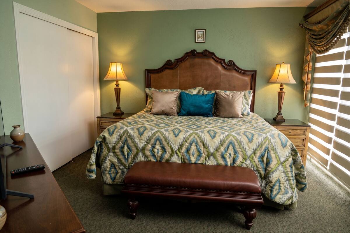 A bedroom with a bed, two bedside tables with lamps, a wooden bench, a dresser, and a door.