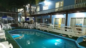 Une piscine entourée de chaises longues et de parasols, elle-même bordée d’un bâtiment doté de nombreuses fenêtres et de balcons.