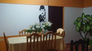 Comedor con una mesa dispuesta para cuatro personas, rodeado de sillas de madera. Un jarrón con flores blancas está sobre la mesa, y una planta en maceta se encuentra a la derecha.