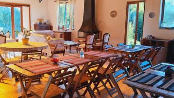 A spacious dining area with multiple wooden tables and chairs, a central round table with a yellow tablecloth, and a large window providing natural light.