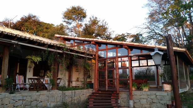 A rustic house with a stone wall, wooden staircase, and a covered porch with chairs and tables. - Chalé na Montanha (Cunha)