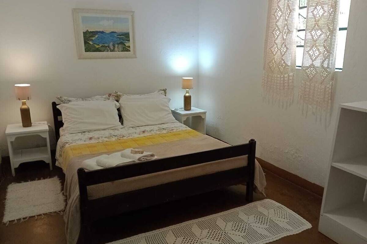 Une chambre à coucher avec un lit, deux tables de chevet, une commode, une fenêtre ornée de rideaux en dentelle et un tableau accroché au mur.