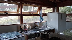 A rustic kitchen with a gas stove, sink, and refrigerator. Large windows provide natural light and views of the outdoors. - Chalé na Montanha (Cunha)