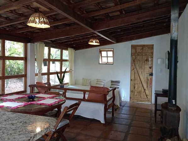 Una habitación rústica con techo de madera, una cama con cabecera de madera, una mesa de comedor con sillas y un ventanal con cortinas.