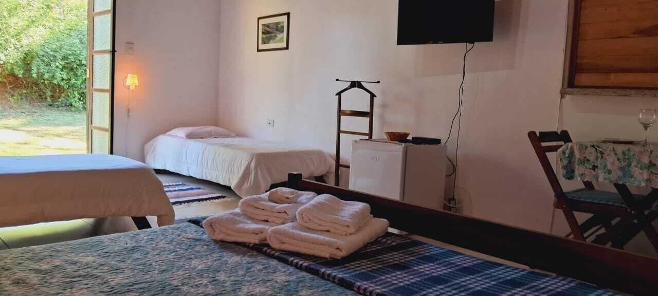 A room with two beds, a table with towels, a television, and a window with a view of greenery.