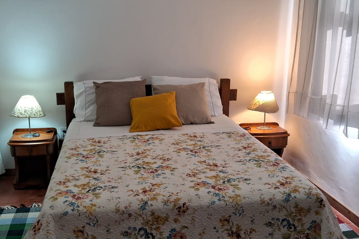 Une chambre d’hôtel avec un lit, deux tables de chevet, une lampe sur chaque table et une fenêtre avec des rideaux légers.