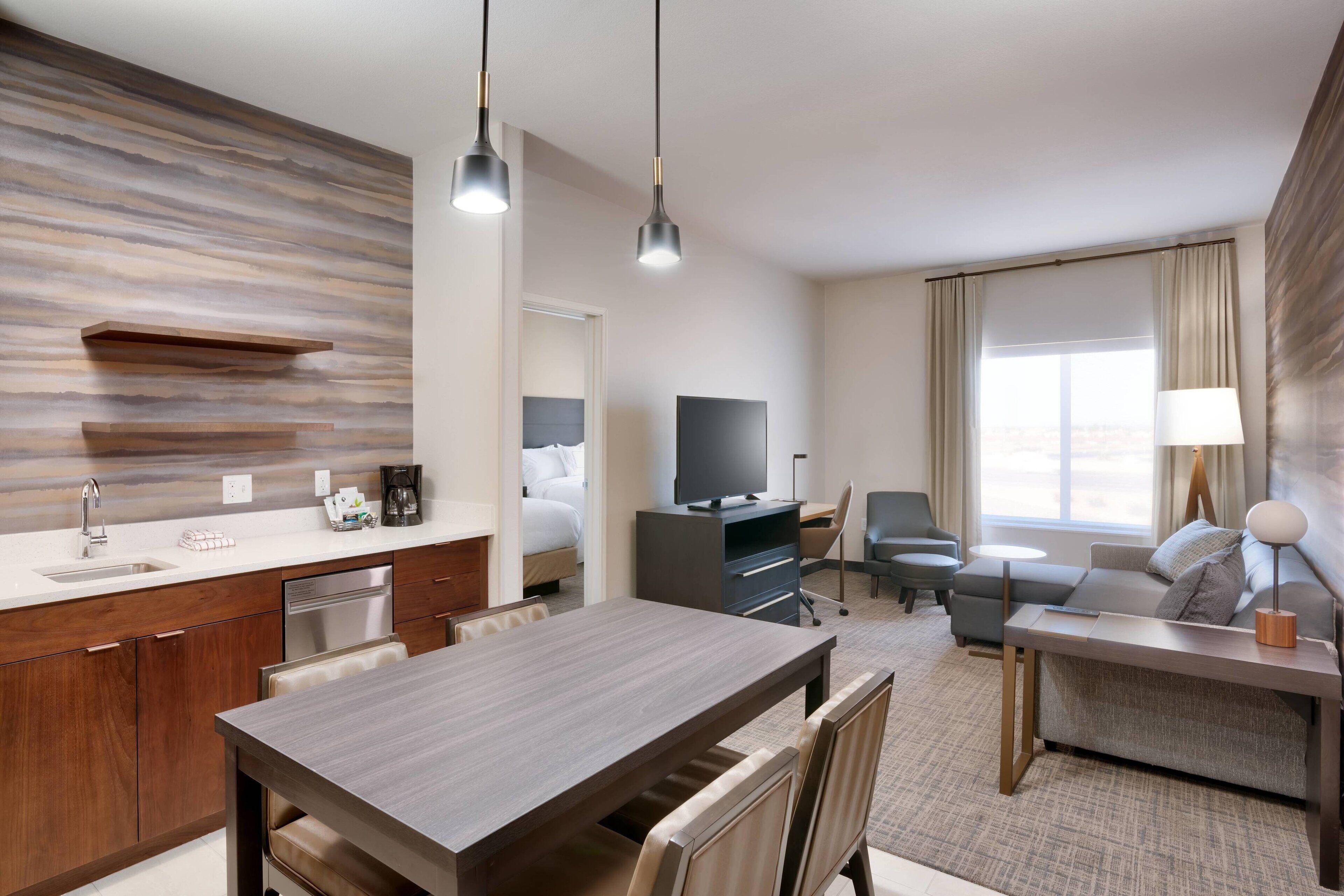 A modern hotel room with a kitchen area, a dining table, a sofa, and a TV.