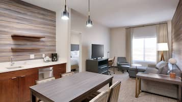 A modern hotel room with a kitchen area, a dining table, a sofa, and a TV.