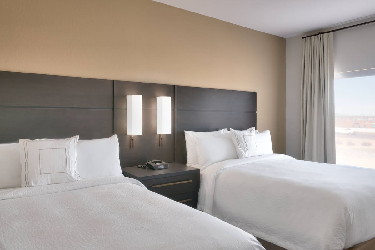 A hotel room with two beds, a nightstand with a phone, and a view of the city through large windows.
