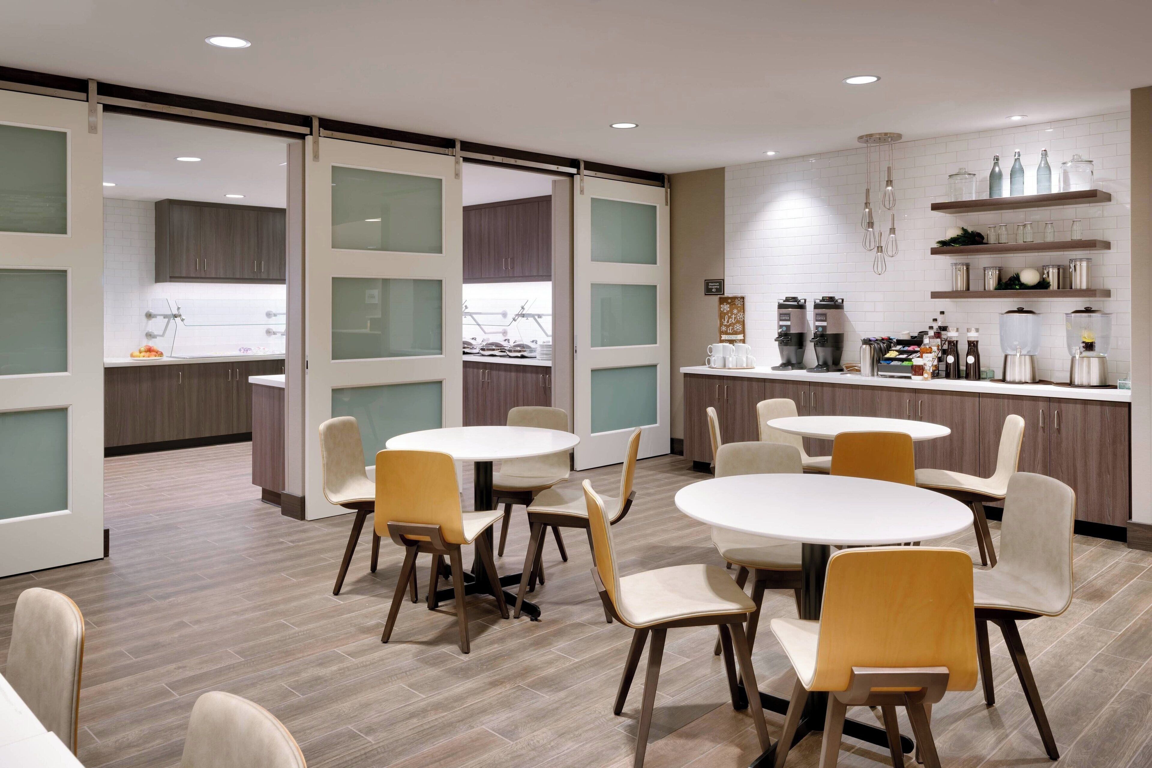 Een moderne eethoek met ronde tafels en stoelen, een keuken op de achtergrond en een schuifdeur naar een andere kamer.