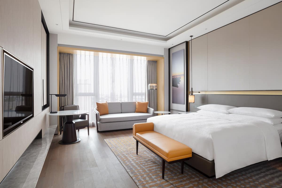 A modern hotel room with a large bed, a sofa, a dining table, and a TV.