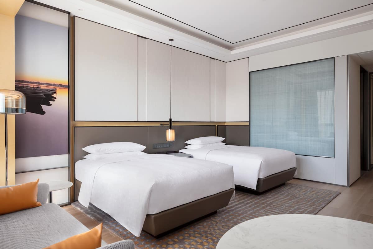 A modern hotel room with two beds, a sofa, a small table, and a large window.