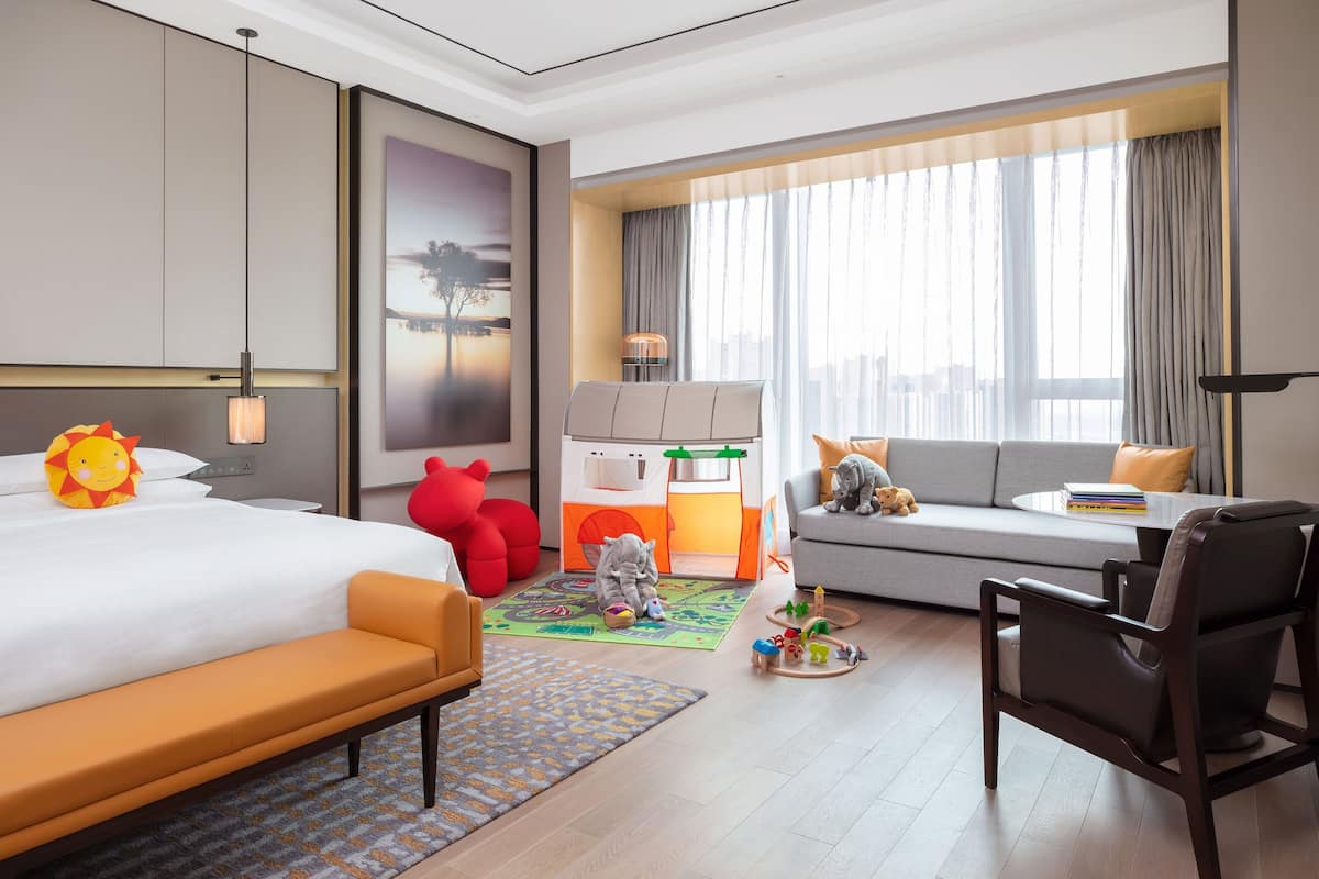 A hotel room with a bed, a sofa, a desk, a chair, a play area with toys, and a large window with curtains.