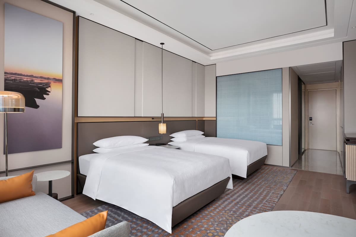 A modern hotel room with two beds, a sofa, a small table, and a large window.