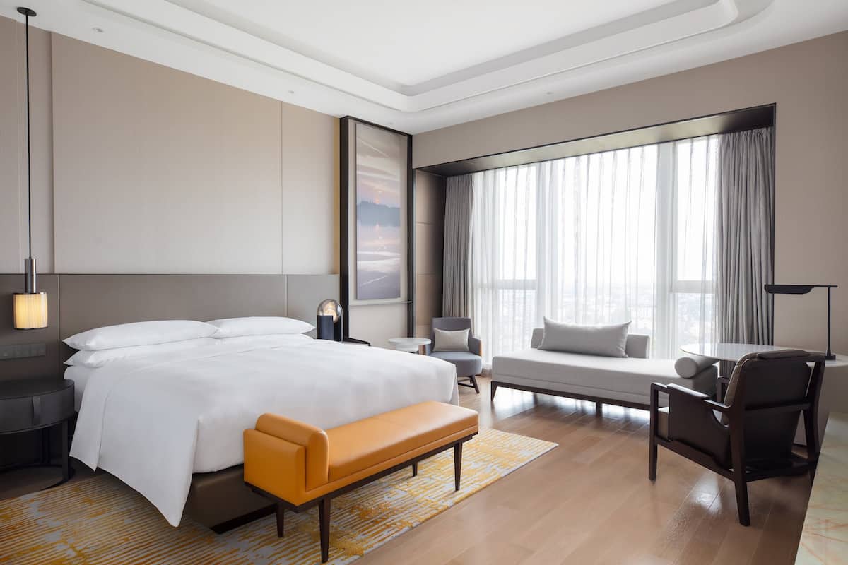 A modern hotel room with a large bed, a seating area with a sofa and armchairs, a small table, and a large window with curtains.