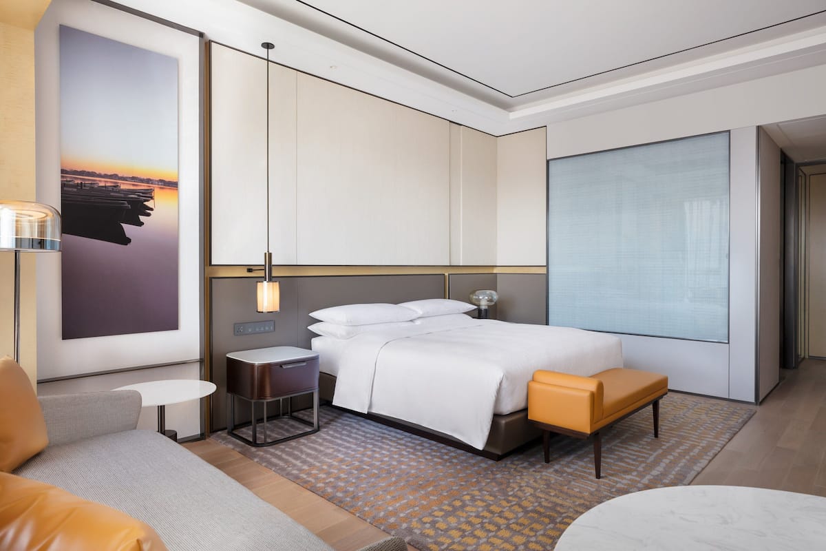A modern hotel room with a large bed, a small table, a bench, and a large window.
