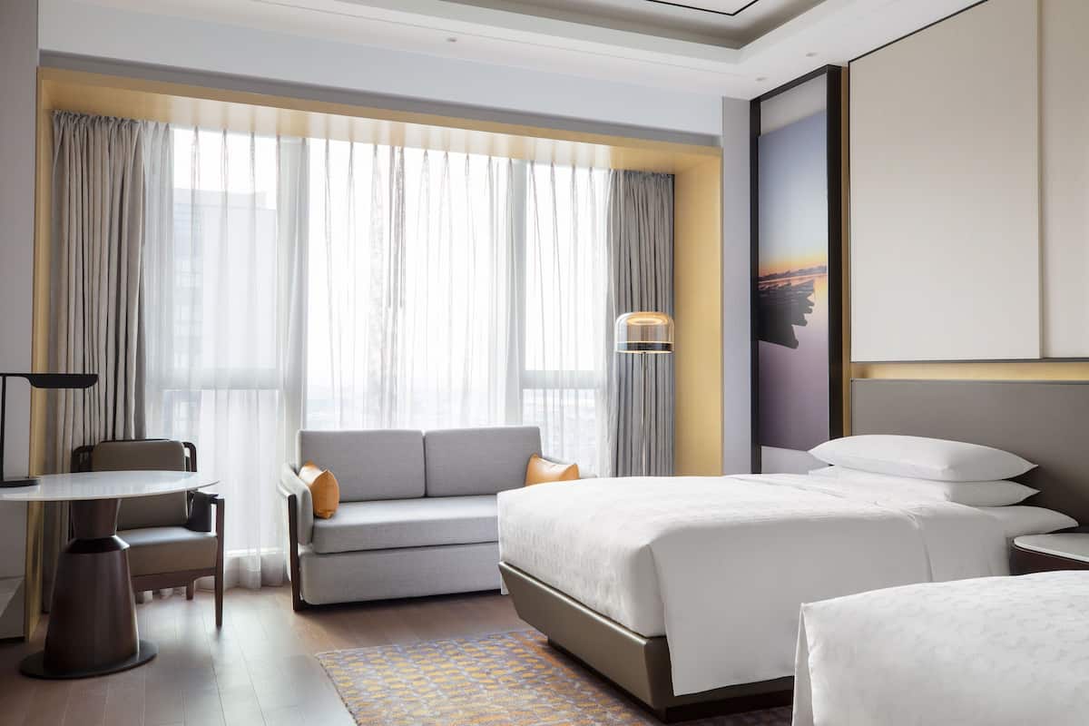 A modern hotel room with a large bed, a sofa, a desk, and a large window with curtains.