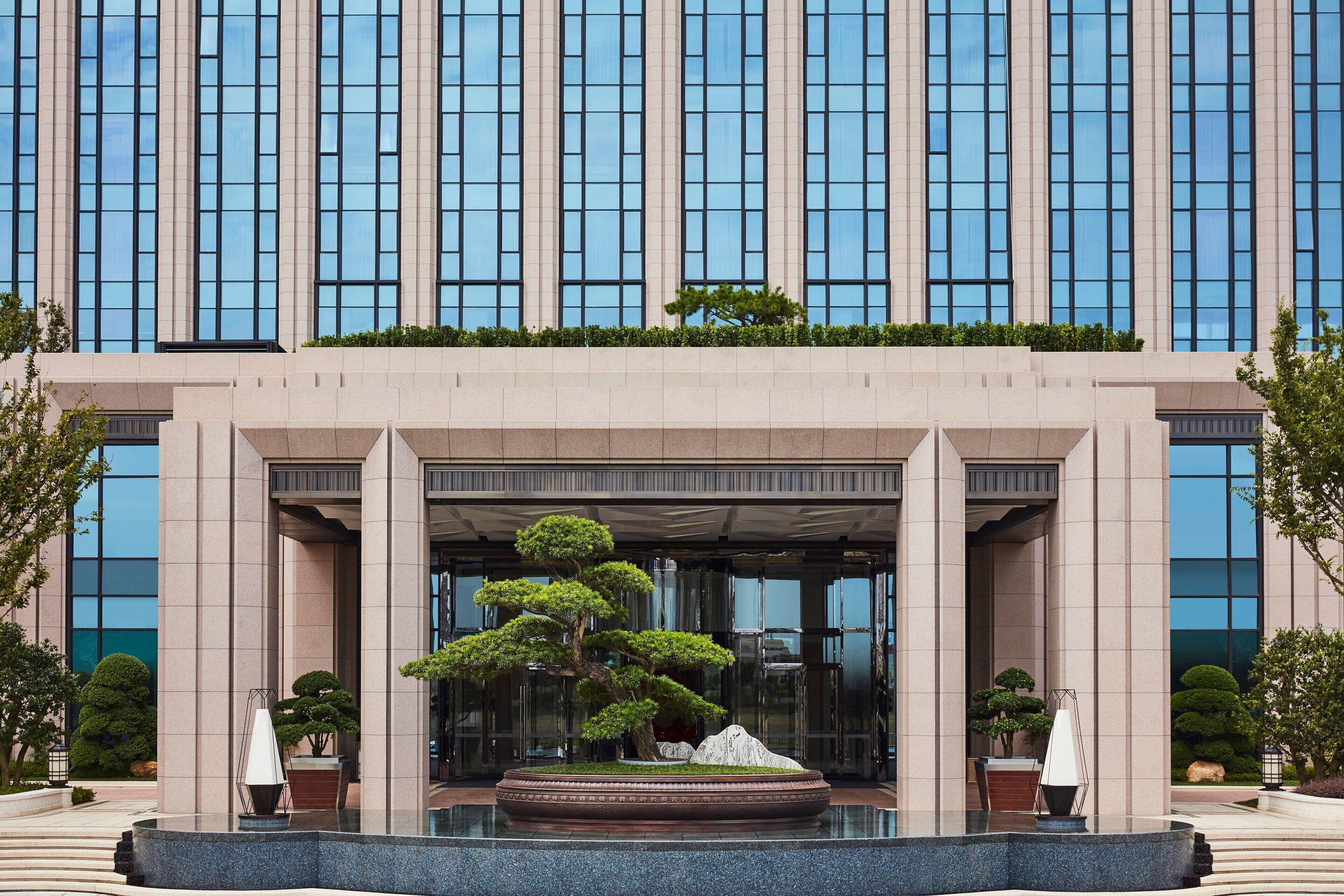 A modern building with a large entrance, a central garden with a bonsai tree, and a fountain.