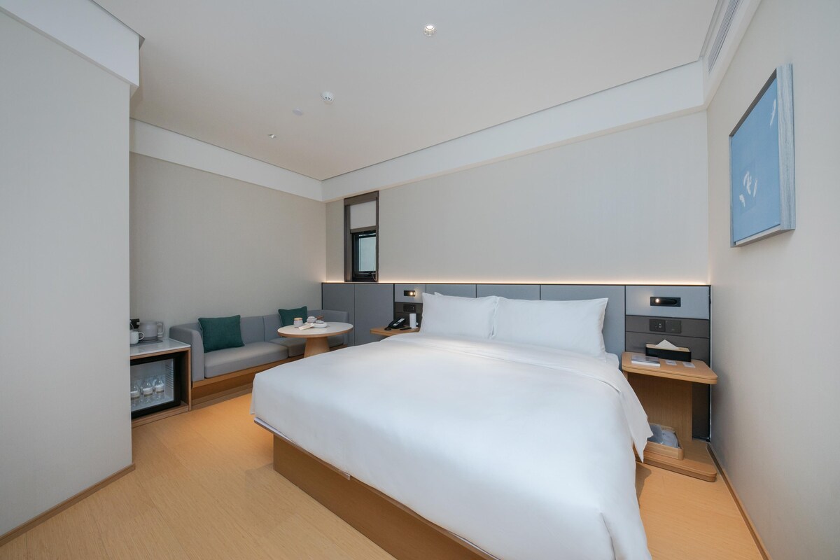 Una habitación de hotel moderna con una cama grande, mesitas de noche, un pequeño rincón de descanso y una mesita auxiliar.