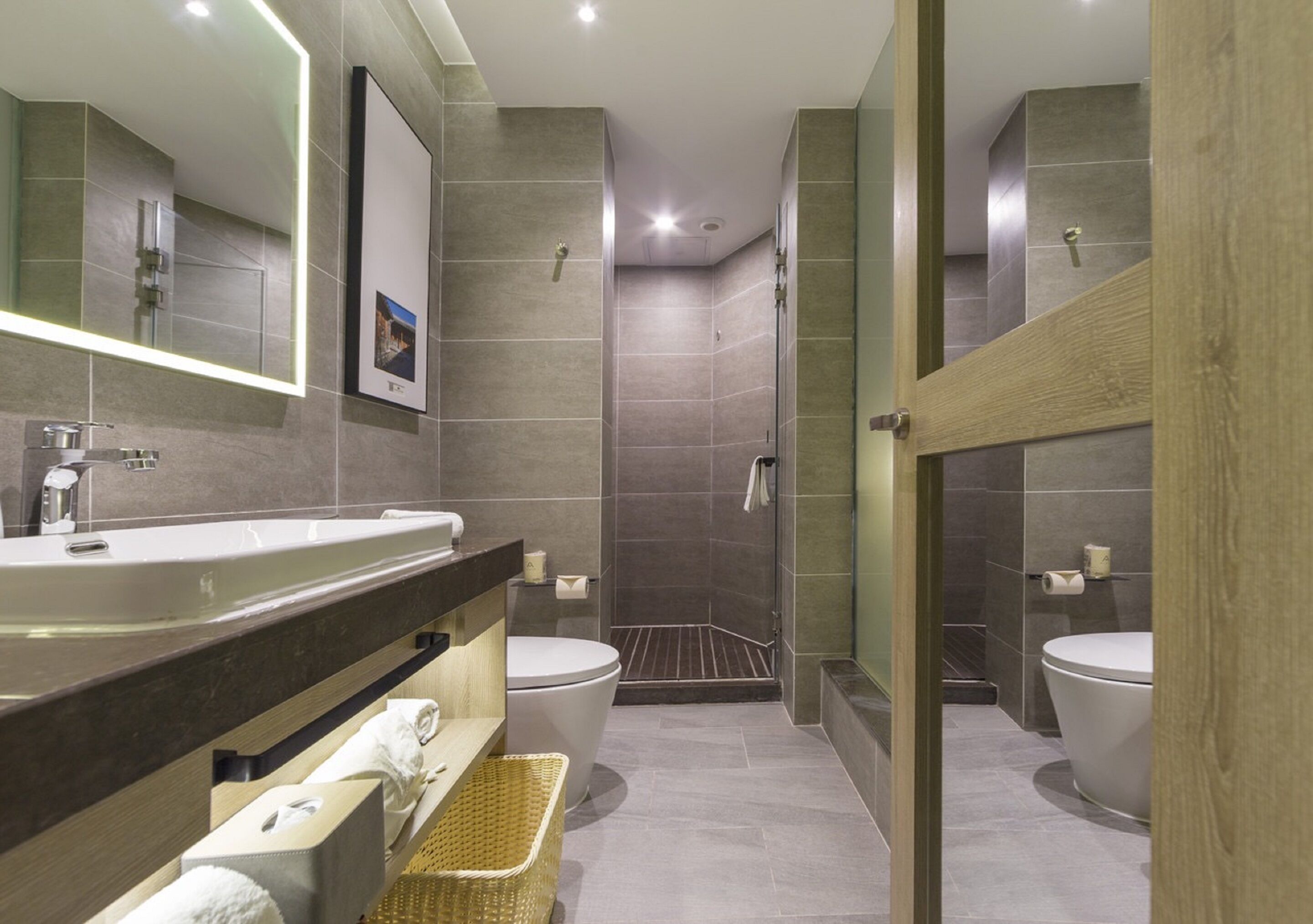 A modern bathroom with a large mirror, a sink, two toilets, and a shower area.